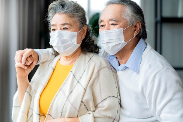 Asian retired elderly couple wearing face masks watch the world through their home window hand hug and support togetherasian people home isolation social distancing stay home ideas concept