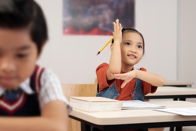 Free photo asian schoolgirl sitting at desk in classroom and lifting up her hand to answer