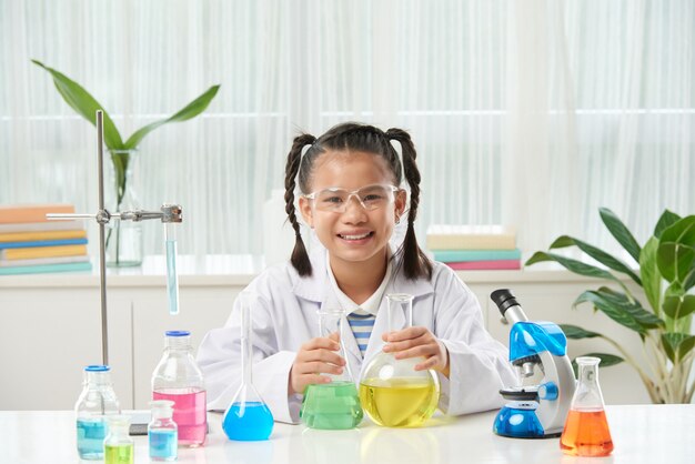 Asian schoolgirl with braids sitting at desk with microscope and vials with colorful liquids