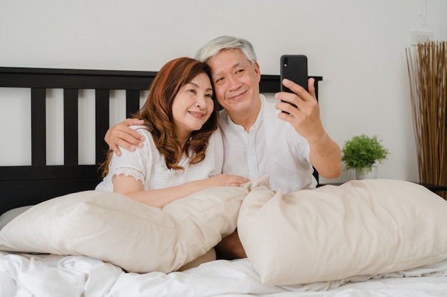 Asian senior couple selfie at home. Asian Senior Chinese grandparents, husband and wife happy using mobile phone selfie after wake up lying on bed in bedroom at home in the morning concept.