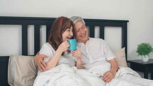 집에서 침대에 아시아 수석 커플입니다. 아침 개념에 집에서 침실에서 침대에 누워있는 동안 일어나 아시아 수석 중국 조부모, 남편과 아내 행복 음료 커피.