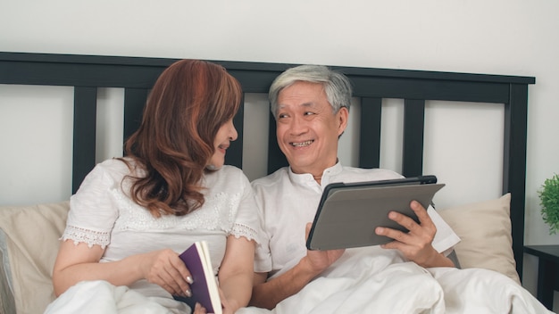 Азиатские старшие пары используя таблетку дома. Азиатские старшие китайские деды, кино просмотра супруга и жена прочитали книгу после бодрствования, лежа на кровати в спальне дома в концепции утра.