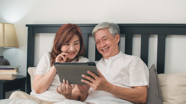 Азиатские старшие пары используя таблетку дома. Азиатские старшие китайские деды, муж и жена счастливые после бодрствования, смотря кино лежа на кровати в спальне дома в концепции утра.