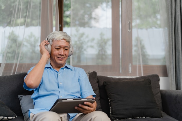 Азиатский старший мужчина расслабиться дома. Азиатские наушники носки более старого мужчины счастливые используя подкаст таблетки слушая пока лежащ на софе в концепции живущей комнаты дома.