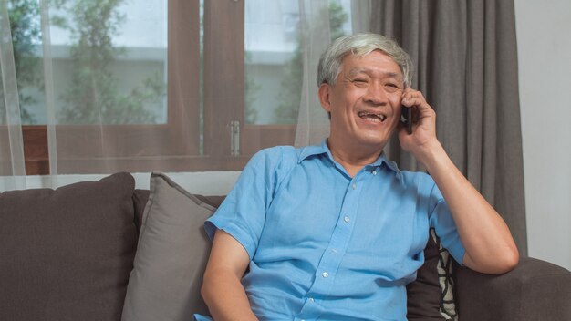 Asian senior man talk on phone at home. Asian senior older Chinese male using mobile phone talking with family grandchild kids while lying on sofa in living room at home concept.