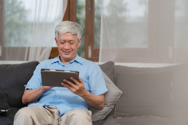 집에서 태블릿을 사용하는 아시아 수석 남자. 집에서 거실에서 소파에 누워있는 동안 인터넷에서 건강을 지키는 방법에 대한 아시아 수석 중국 남성 검색 정보 개념.