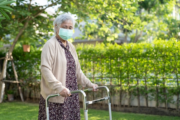 アジア​の​高齢者​または​高齢​の​老​婦人​女性​は​、​歩行器​を​持って​歩き​、​安全​感染​を​防ぐ​ために​フェイス​マスク​を​着用します​。