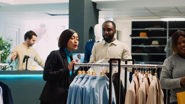 タブレットを使って店舗の在庫を調べ、ハンガーやラックにあるすべてのフォーマルウェアを分析するアジア人の店員。衣料品店で衣類の在庫を調べ、製品を調べる若い労働者。