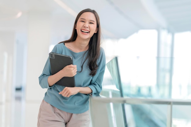 Asian smiling cheerful female digital nomad hand hold tablet device look at camera portrait shothappiness smiling asia woman standing in office college corridor with positive smiling attitude