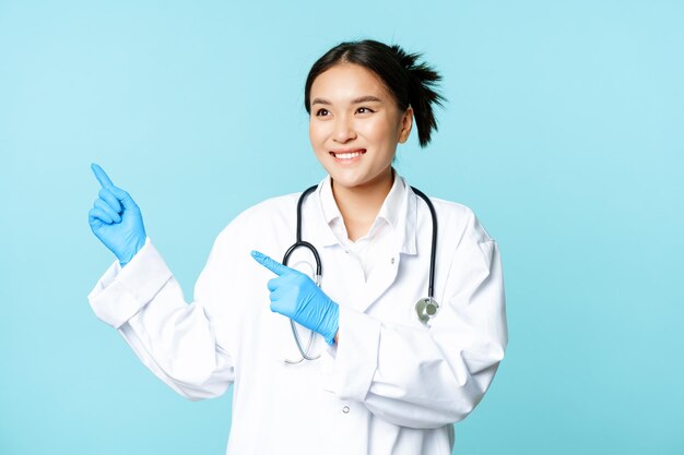Asian smiling doctor medical healthcare worker pointing fingers left looking pleased at advertisemen...