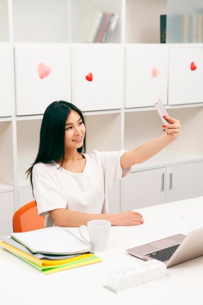 Asian student photographing herself in library Brunette lady holding mobile or smart phone in front of her while studying