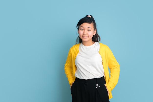 Asian teenager's portrait isolated on blue studio background. Beautiful female brunette model with long hair. Concept of human emotions, facial expression, sales, ad. Posing, looks confident.