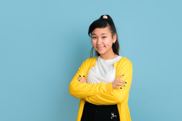 Asian teenager's portrait isolated on blue studio background. Beautiful female brunette model with long hair. Concept of human emotions, facial expression, sales, ad. Posing with hands crossed.