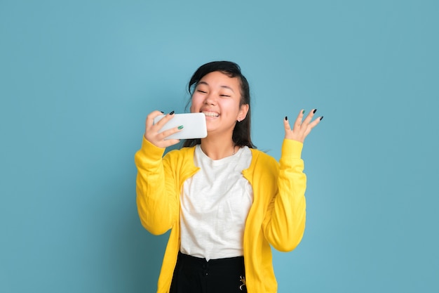 Asian teenager's portrait isolated on blue studio background. Beautiful female brunette model with long hair. Concept of human emotions, facial expression, sales, ad. Using phone, smiling, happy win.