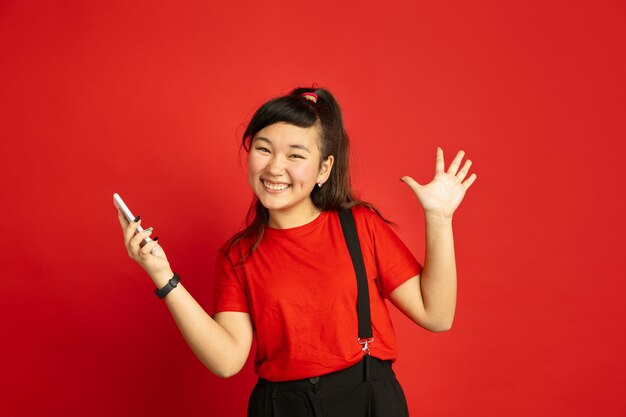 Asian teenager's portrait isolated on red studio background. Beautiful female brunette model in casual style. Concept of human emotions, facial expression, sales, ad. Happy, holding smartphone.