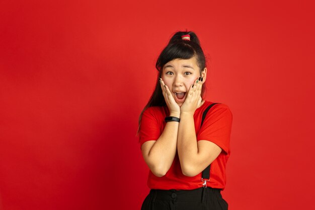 Asian teenager's portrait isolated on red studio background. Beautiful female brunette model with long hair in casual. Concept of human emotions, facial expression, sales, ad. Astonished, shocked.