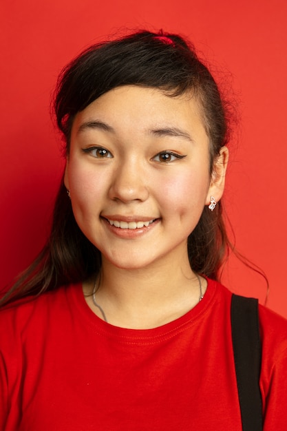 Asian teenager's portrait isolated on red studio background. Beautiful female brunette model with long hair in casual style. Concept of human emotions, facial expression, sales, ad. Smiling cute.