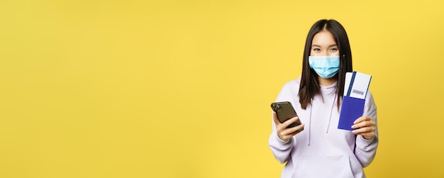 Asian tourist girl in face mask holding mobile phone showing passport and flight tickets booking hot