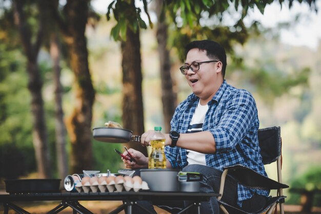 Asian traveler man glasses frying a tasty fried egg in a hot pan at the campsite Outdoor cooking traveling camping lifestyle concept