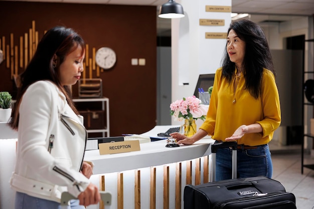 アジアの旅行者が休暇中に居心地の良いホテルのロビーに到着し、受付係からのサービスを期待しています。予約をサポートするコンシェルジュを待つ新しいリゾートゲスト