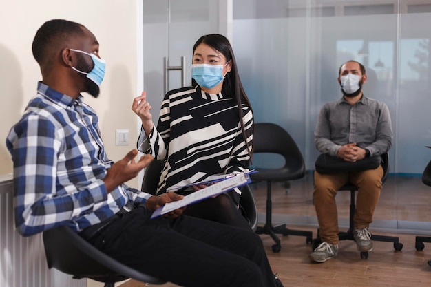 Asian unemployed woman and african american man sitting in lobby area of financial company while talking about job oportunity. Diverse people in waiting room of agency talking about employment chance.