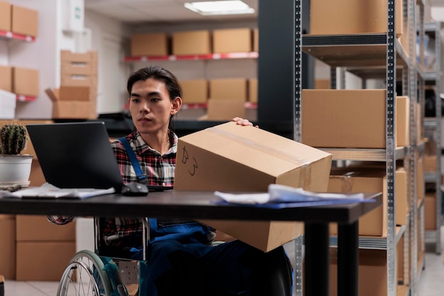 Free photo asian wheelchair user managing parcel delivery in postal service warehouse. storehouse shipment operator with disability holding cardboard box with customer order and planning shipping route on laptop