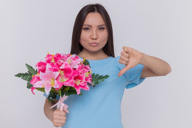 Азиатская женщина в синем платье держит букет цветов, недовольно глядя вперед, показывая большой палец вниз, празднуя международный женский день, стоя над белой стеной