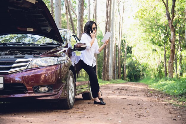 Asian woman calling repairman or insurance staff to fix a car engine problem on a local road 