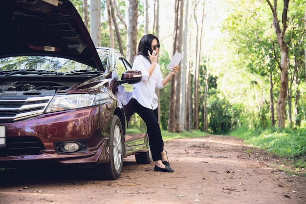 Asian woman calling repairman or insurance staff to fix a car engine problem on a local road 