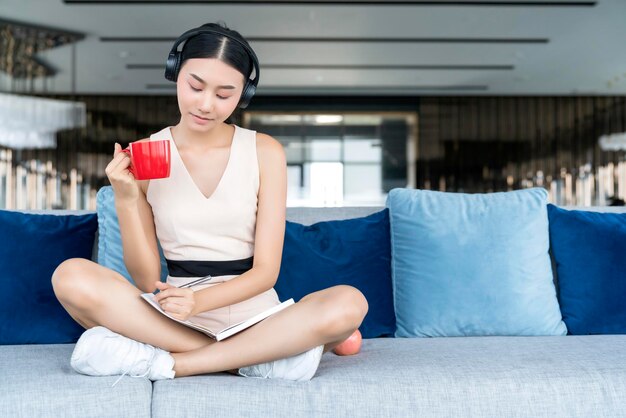 캐주얼한 작업복을 입은 아시아 여성은 거실 배경의 소파에서 커피와 잡지와 함께 휴식을 취합니다.
