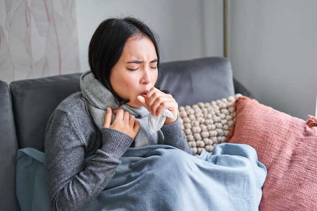 Asian woman coughing feeling sick sitting under blanket with scard in cold apartment saving on heati