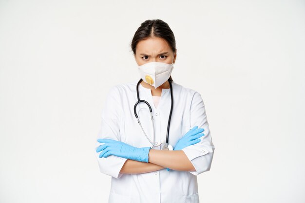 Asian woman doctor in medical face respirator and rubber gloves, looking angry at camera, cross arms on chest, standing in healthcare worker uniform, white background.