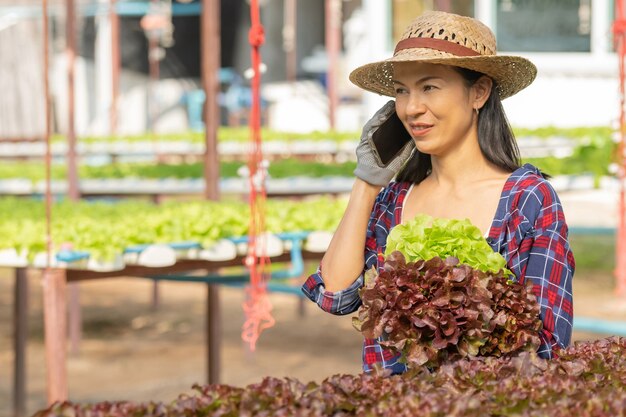 野菜の水耕栽培でモバイルを使って幸せに働くアジアの女性農家。グリーンハウス農場で、グリーンサラダ野菜の品質を笑顔でチェックする女性農家のポートレート。