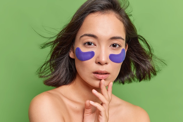 Asian woman has dark hair floating in air applies blue hydrogel patches under eyes undergoes skin care