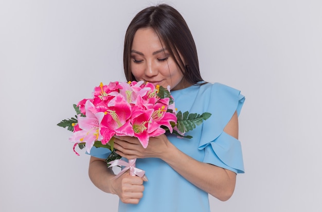 흰색 벽 위에 서있는 국제 여성의 날을 축하하는 행복하고 쾌활한 꽃의 꽃다발을 들고 아시아 여자