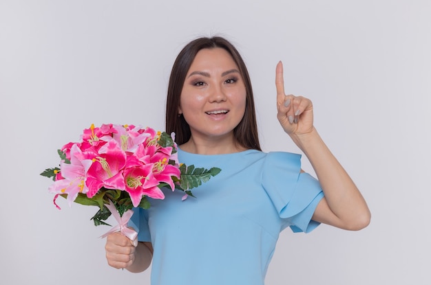 Азиатская женщина, держащая букет цветов, выглядит счастливой и веселой, улыбаясь показывая указательный палец, празднуя международный женский день, стоя над белой стеной