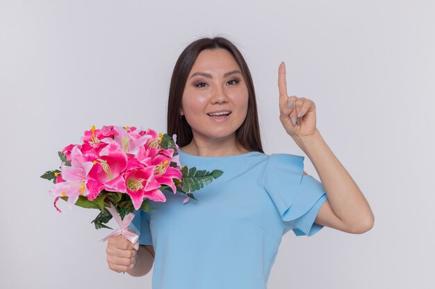 Asian woman holding bouquet of flowers looking happy and cheerful smiling showing index finger celebrating international women's day standing over white wall