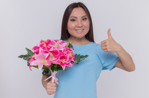 Asian woman holding bouquet of flowers looking happy and cheerful smiling showing thumb up celebrating international women's day standing over white wall