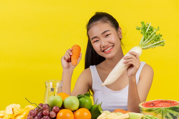 Азиатская женщина, держащая морковку правой рукой Держите редьку левой рукой и на столе много фруктов.