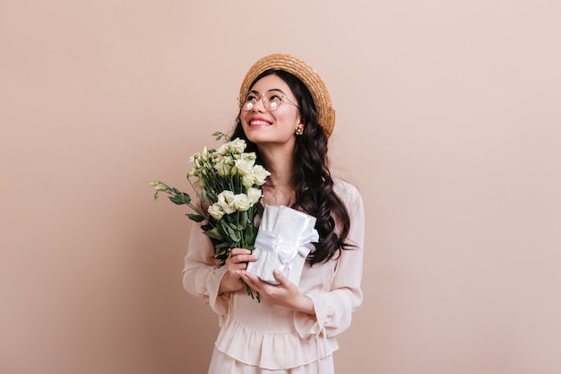 花とプレゼントを持っているアジアの女性。ベージュで分離されたトルコギキョウの花束を持つインスピレーションを得た日本人女性のスタジオショット。