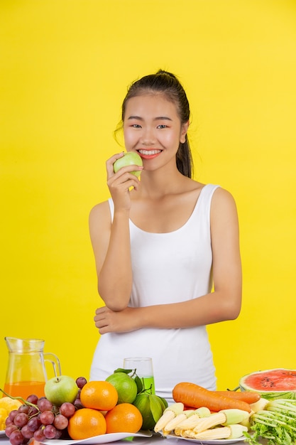 An Asian woman holding a green apple with her right hand, and on the table there are many fruits.