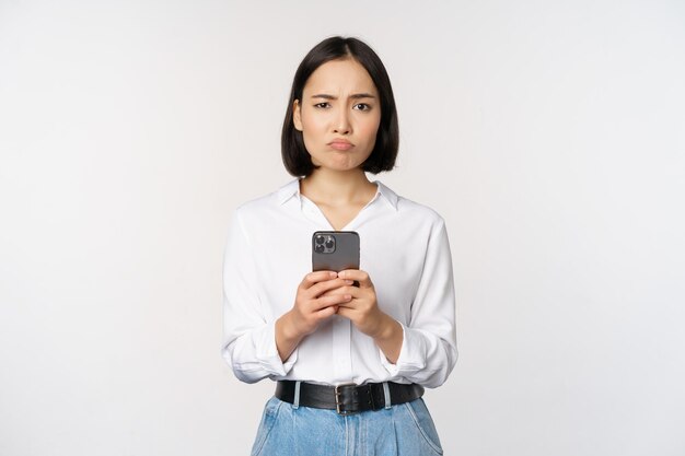 스마트폰을 들고 의심스러운 표정으로 바라보는 아시아 여성은 흰색 배경에 반대하는 휴대전화 앱에 실망했다