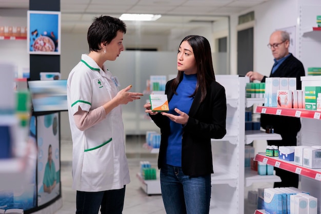 Asian woman holding vitamin package and listening to pharmacist explaining ascorbic acid dosage instruction. Drugstore customer buying food supplement, talking with pharmacy assistant in aisle