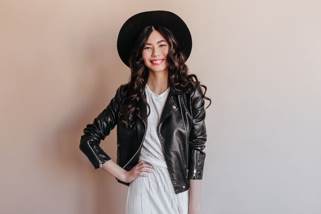Asian woman in leather jacket standing with hand on hip. Studio shot of smiling chinese woman in hat isolated on beige background.