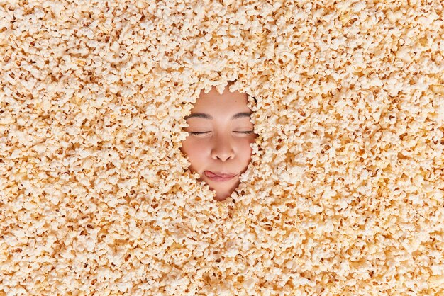 Asian woman licks lips keeps eyes closed imagines eating appetizing snack drowned in delicious sweet popcorn going to watch movie with friends. Overhead shot. Yummy air popped corn