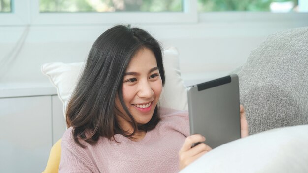 アジアの女性は音楽を聴くとタブレットを使用して、女性は自宅のリビングルームの家のソファーに横たわってリラックスした時間を使用します。幸せな女性は、ヘッドフォンのコンセプトで音楽を聴きます。