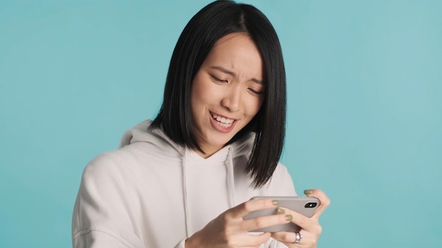 Asian woman looking nervous emotionally playing on smartphone over blue background