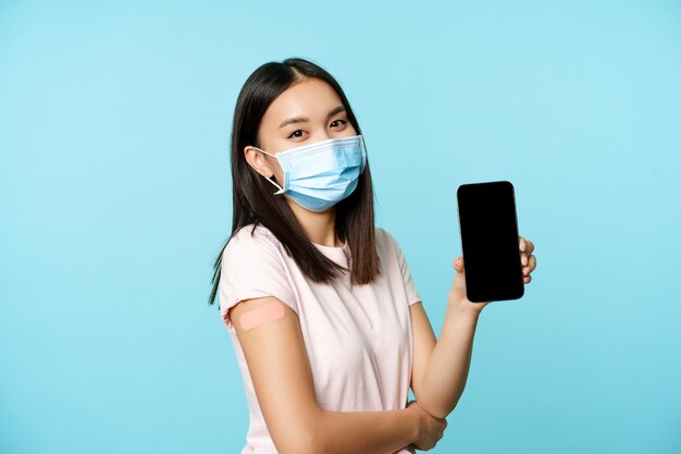 의료용 얼굴 마스크를 쓴 아시아 여성, 코비드-19 백신 후 패치가 있는 팔, 앱 인터페이스가 있는 스마트폰 화면, 예방 접종 증명서 개념, 건강 여권