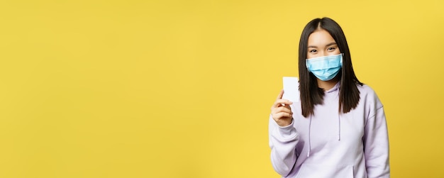 Free photo asian woman in medical face mask showing credit card pass standing over yellow background