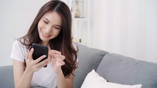 Азиатская женщина играя smartphone пока лежащ на домашней софе в ее живущей комнате.